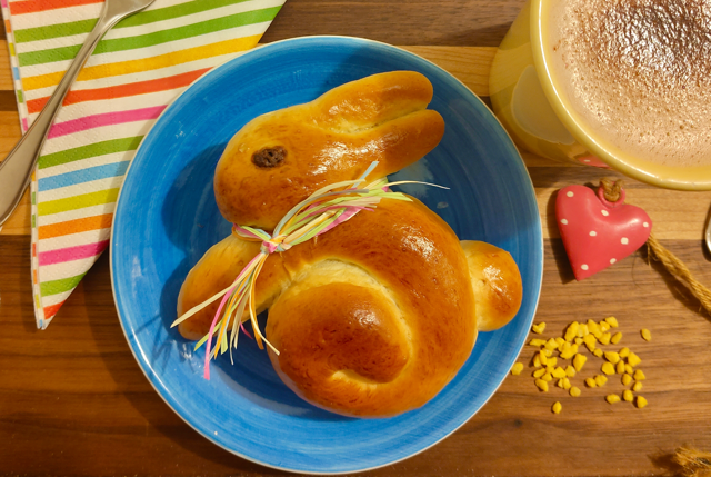 Hefeosterhäschen mit buntem Band, serviert auf einem blauen Teller, neben einer Tasse.