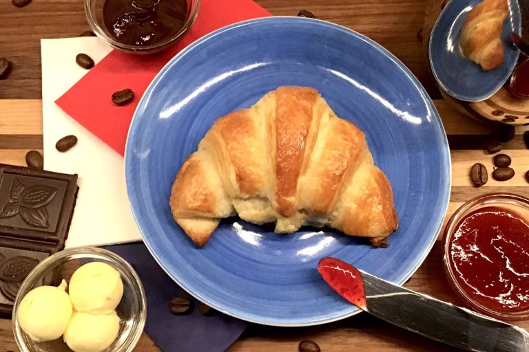 Ein Croissant auf einem blauen Teller, umgeben von Marmelade, Schokolade und Butterkugeln.