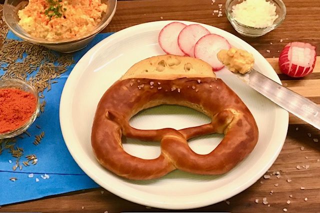 Brezel mit Senf, Radieschen und Gewürzen auf einem weißen Teller.